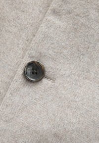Lys grå, tekstureret uldstof med en mørkegrå knap med fire huller, syet på en klap søm, hvilket tilføjer en raffineret detalje.
