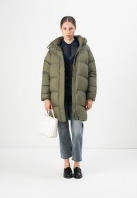 Olijfgroene pufferjas met capuchon, met een ritssluiting aan de voorkant, doorstikte design, gedragen met donkere wijde jeans en een witte handtas.