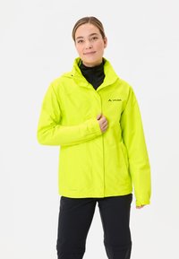 Veste imperméable jaune avec capuche, coupe ajustée et poignets réglables. Logo de la marque visible sur la poitrine droite. Portez sur un haut noir.