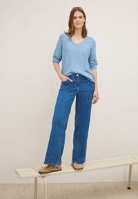 Helles blaues V-Ausschnitt-Sweatshirt mit hochgerollten Ärmeln, kombiniert mit hochtaillierten, weit geschnittenen blauen Jeans. Das Model steht auf einer Metallbank und trägt beigen Slipper.