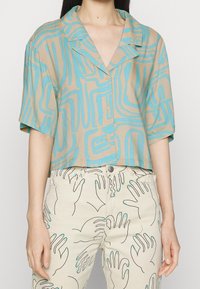 Krótki rękaw, krótka brązowa koszula z turkusowymi, abstrakcyjnymi liniami. Posiada kołnierzyk i zapięcie na guziki. Noszona z lekkimi spodniami z wzorami rąk.