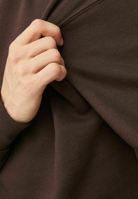 Bruine katoenen sweater met ribgebreide manchetten, die wordt getrokken om de textuur te laten zien; close-up van een hand die de stof vasthoudt.