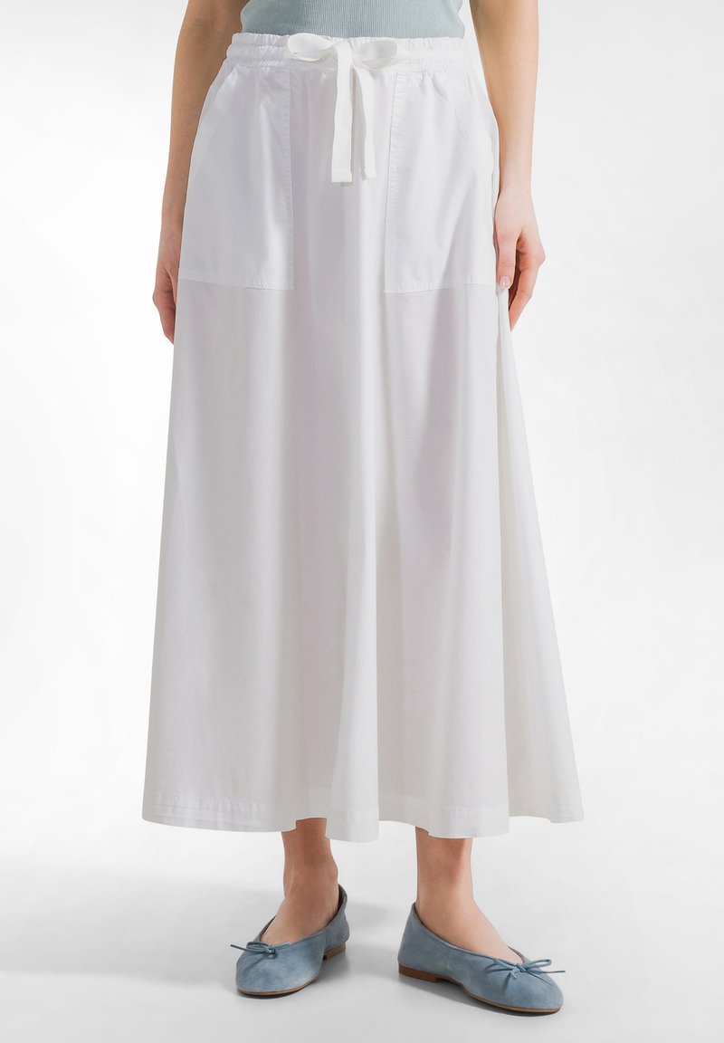 Kvinde iført en lang hvid nederdel med forlommer og snøre i taljen, kombineret med lyseblå ballet flats med sløjfer.