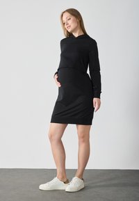 Robe de maternité noire avec un crop-top à capuche, des manches longues et une coupe ajustée à la taille. Associée à des baskets blanches pour un look décontracté.