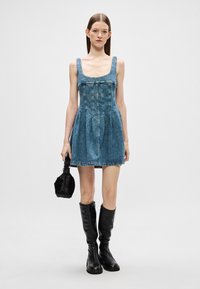 Jeans φόρεμα με τετράγωνη λαιμόκοψη, εφαρμοστό μπούστο και φουσκωτή φούστα. Φορεμένο με μαύρες μπότες μέχρι το γόνατο και κρατώντας μια μικρή μαύρη τσάντα.