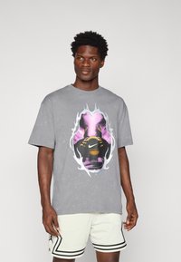 Grå bomulds T-shirt med et stort, farverigt grafisk motiv i midten. Designet inkluderer flammer og et logo. Modellen har lette shorts på.