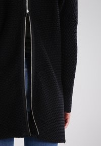 Schwarz strukturierter Mantel mit einem teilweise offenen vertikalen silbernen Reißverschluss auf dem Rücken, getragen über blauen Jeans, mit einer sichtbaren Hand an der Seite.