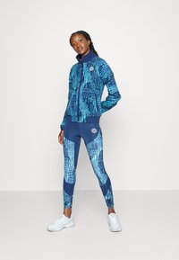 Blauw gemarmerd sportieve outfit bestaande uit een ritsjack met geometrische ontwerpen en bijpassende leggings. Witte sneakers maken de look af.