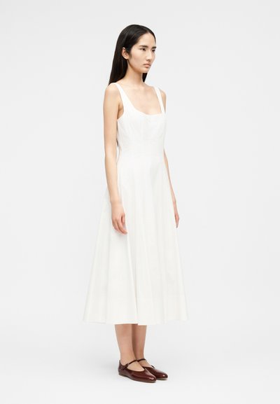 Νέα γυναίκα με μακριά σκούρα μαλλιά, ντυμένη με μίντι λευκό φόρεμα χωρίς μανίκια και καφέ παπούτσια Mary Jane, στέκεται μπροστά σε απλό ανοιχτό φόντο.