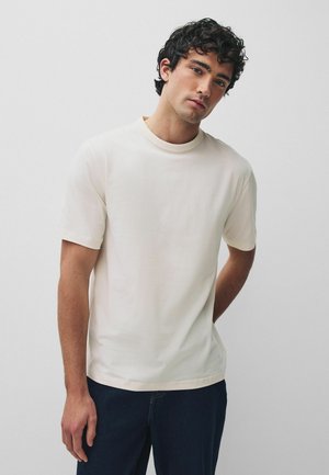 Ung mand med krøllet hår iført en ensfarvet creme kortærmet t-shirt og mørke bukser, stående mod en lysegrå baggrund.