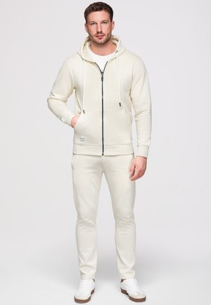 Mann steht mit der Hand in der Tasche, trägt einen off-white Hoodie, passende Jogginghosen und weiße Sneakers vor einem einfarbigen hellen Hintergrund.