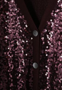 Mélykádvári színű flitteres kardigán kör alakú rózsaszín flitterekkel. Elöl két fekete gombbal. Simább textúrájú, fényes díszítésekkel.