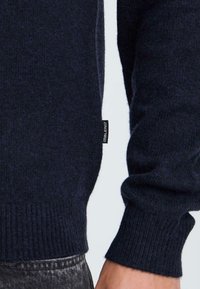 Dunkelblauer gestrickter Pullover mit gerippten Bündchen und Saum, der ein kleines schwarzes Etikett an der Seite hat. Das Material wirkt weich und strukturiert.