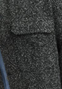 Un tissu texturé noir et gris présente une poche poitrine et des coutures visibles. La surface affiche un motif tissé avec des fibres variées.