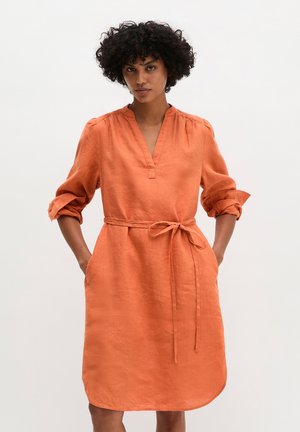 Frau mit lockigem Haar trägt ein knielanges orangenes Kleid mit hochgekrempelten Ärmeln und einem gebundenen Taillengürtel, steht vor einem schlichten Hintergrund.