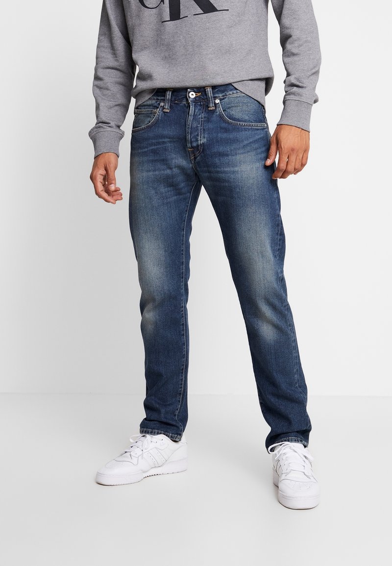 Homme portant un jean bleu délavé, un sweat gris avec de grandes lettres noires et des baskets blanches, debout devant un fond blanc.