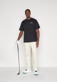 Μαύρο βαμβακερό μπλουζάκι με μικρό λογότυπο, κρέμα παντελόνι, λευκά παπούτσια γκολφ με μπλε λεπτομέρειες, κρατώντας ένα γκολφ, στέκεται σε ουδέτερο φόντο.