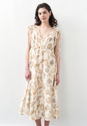 Frau trägt ein ärmelloses, cremefarbenes Midikleid mit beigen und braunen Muschel- und Seesternmustern, steht vor einem schlichten hellen Hintergrund.