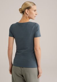 Tricou scurt cu mânecă închis la culoare teal, cu guler rotund. Textură moale și netedă. Design ajustat care conturează corpul. Purtați cu pantaloni gri deschis.