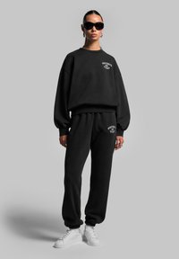 Schwarzer, übergroßer Pullover und passende Jogginghosen aus einem weichen Baumwollmix. Der Pullover hat ein Logo auf der Vorderseite; die Jogginghosen haben ein Logo auf der Rückseite. Weiße Sneaker.