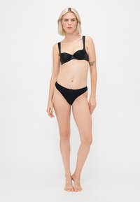Conjunto de lingerie preta com um sutiã liso e almofadado e uma cueca em estilo biquíni a condizer, adornado com um delicado laço, colocado sobre um fundo simples.
