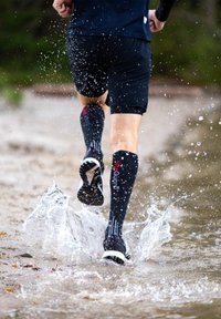 Černé sportovní šortky a kompresní ponožky do lýtek s červeným logem, rozstřikující vodu při běhu v mělké vodě na pláži.