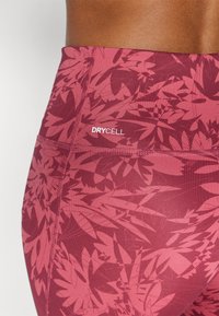 Detailní záběr na vínové legíny se květinovým vzorem, vyrobené z materiálu odvádějícího vlhkost, se švy a logem "DRYCELL" na pase.