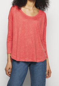 Koralli pitkähihaine paita, jossa on pyöreä kaula-aukko, rento malli, kevyet materiaalit ja sivuhalkiot; yhdistetty sinisiin farkkuihin.