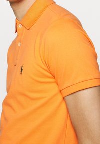 Orange polo skjorta, blandning av bomull, kortärmad, ribbad krage, tvåknappars placket, med en liten grön broderad logotyp på bröstet.