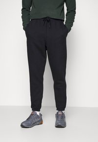 Osoba v černých joggingových kalhotách, tmavě zeleném dlouhém rukávu, šedých a oranžových běžeckých botách, stojící proti bílému pozadí.