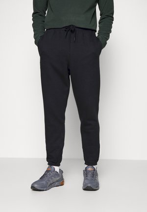 Person klädd i svarta joggingsbyxor, en mörkgrön långärmad tröja och grå och orange löparskor, stående mot en vit bakgrund.