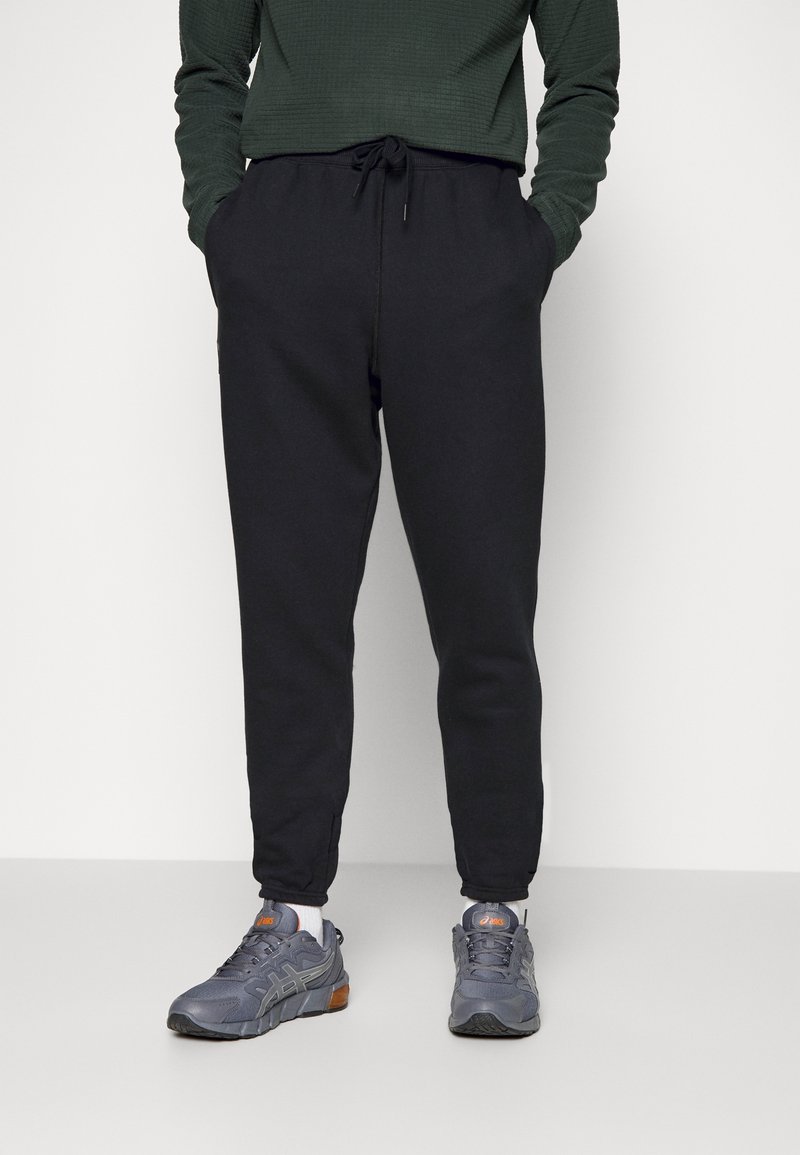 Osoba v černých joggingových kalhotách, tmavě zeleném dlouhém rukávu, šedých a oranžových běžeckých botách, stojící proti bílému pozadí.