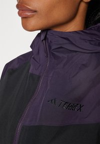 Lilla og svart lett jakke med høyt krage og glidelås, med brodert "TERREX"-logo på brystet. Jevn tekstur.
