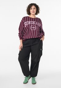 Svitek s črtami Bordeaux z belim napisom, v kombinaciji s črnimi cargo hlačami in zelenimi supergami. Ima ohlapen kroj in elastične manšete.