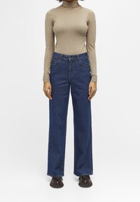Femme portant un pull à col roulé beige, un jean bleu à taille haute et jambes larges, et des chaussures marron foncé, debout devant un fond blanc uni.