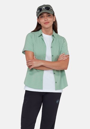 Kvinde iført en grøn ternet kortærmet skjorte over en hvid T-shirt, sorte bukser, grå kasket med solbriller på toppen, krydsede arme.