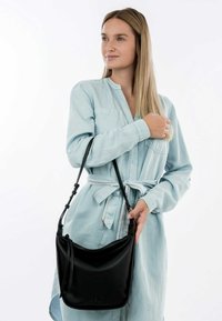 Sac à bandoulière en cuir noir avec une forme courbée, une bandoulière réglable et une marque discrète. Porté avec une robe chemise bleu clair.