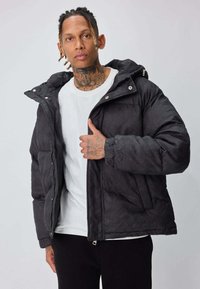 Veste noire matelassée avec un motif chevrons, dotée d'une capuche et de boutons-pression. Portée par-dessus un t-shirt blanc, avec une coupe décontractée.