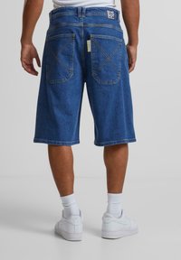 Tummansinisiä denim-Shortseja, joissa on väljä malli. Takataskuissa on viisto sauma ja vyötärön yläpuolella on brändin tarra. Valkoiset tennarit näkyvät.