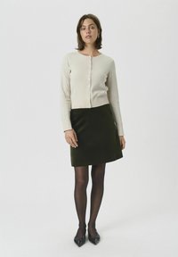 Romige cardigan met knoopsluiting, gecombineerd met een donkergroene rok. Het model draagt zwarte panty's en puntige zwarte schoenen. Eenvoudig en klassiek ontwerp.