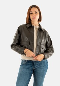 Giacca in pelle lucida e scura con colletto, chiusura a bottoni e due tasche frontali, abbinata a un maglione chiaro e jeans blu.