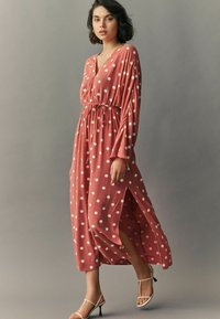 Rochie lungă, de culoare corai, cu buline albe, de tip V, talie strânsă, cu mâneci lungi. Prezintă fante laterale și o siluetă fluidă.