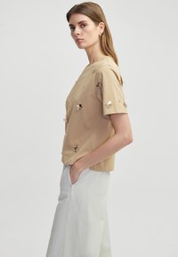 Beige top s krátkým rukávem a dekorativními kovovými detaily, kulatým výstřihem a volným střihem, v kombinaci se světle šedými širokými kalhotami.