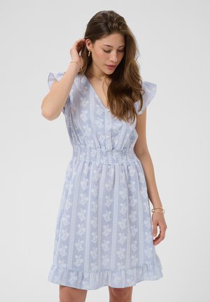 Donna con lunghi capelli castani, che indossa un abito floreale azzurro chiaro con maniche e orlo arricciati, guarda verso il basso e si tocca i capelli.