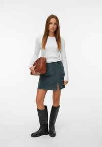 Jupe mini en suède verte avec une fente sur le côté, associée à une chemise blanche à manches longues et un sac à main en cuir marron. Bottes noires jusqu'aux genoux.