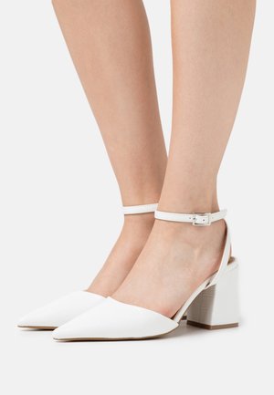 Talons aiguilles blanches à bride de cheville avec un motif texturé rappelant les reptiliens, bout pointu et talon épais et solide. Design à dos ouvert.