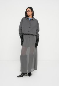 Γκρι κοντό σακάκι με κουμπιά, συνδυασμένο με διαφανή γκρι μακριά φούστα. Μακριά μαύρα γάντια και μυτερά μαύρα τακούνια ολοκληρώνουν το σύνολο.