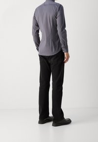 Camisa gris de botones con mangas largas, textura suave, diseño ajustado, combinada con jeans negros y calzado oscuro, vista desde atrás.