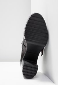 Chaussure en cuir noir avec une semelle en caoutchouc rainurée, dotée d'un talon épais et de détails décoratifs sur la bride. La chaussure est positionnée en diagonale.