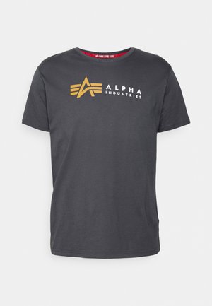 T-shirt gris en coton, avec un col rond. L'avant affiche un logo en or et blanc avec le texte "ALPHA INDUSTRIES."